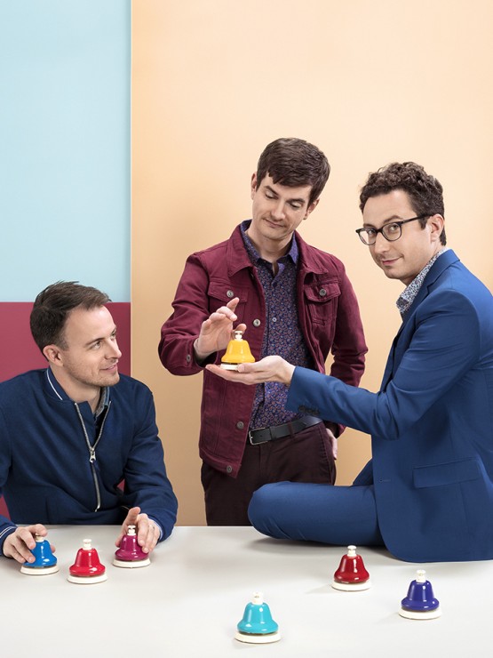 against a muted colorblock background, the four white men of a percussion ensemble hold colorful bells and other percussion instruments