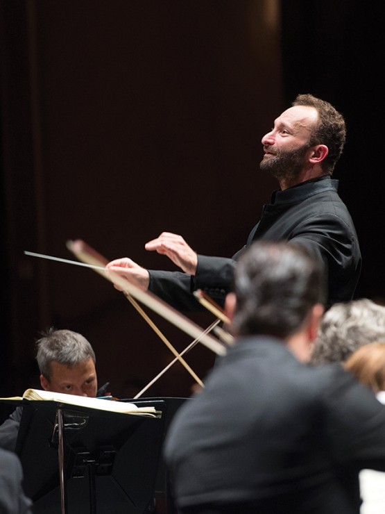 a conductor, focusing intently, pauses. he extends his baton flat and joins the thumb and forefinger of his other hand.