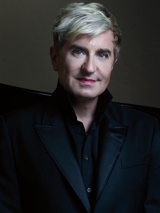 a platinum haired white man, all in black, sits on a dark sofa, his face illuminated