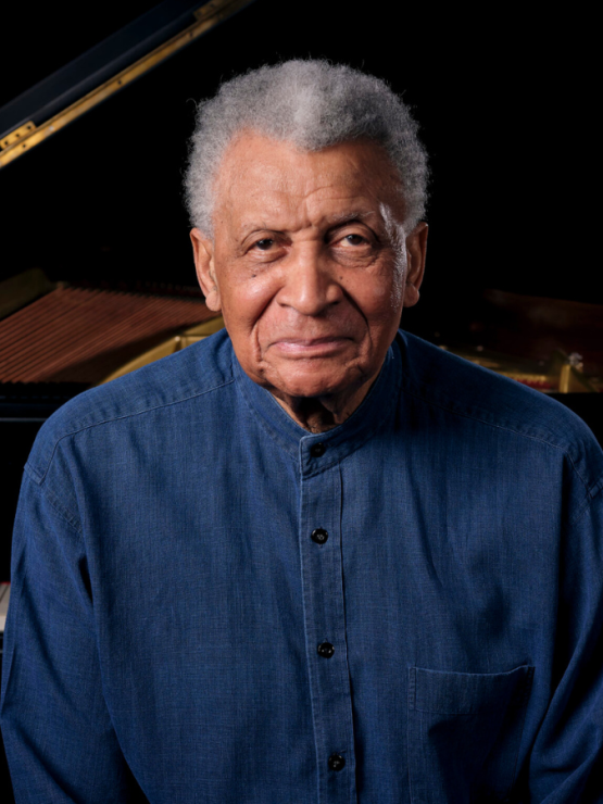 a multiracial man in his late 80s with medium brown skin and short gray and white hair stands with his back to the keyboard of a grand piano
