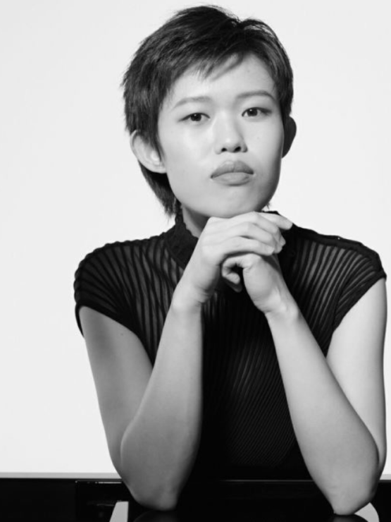 a chinese american woman in her 20s, with short hair in a pixie cut. she rests her elbows on a piano, her hands clasped in front of her face