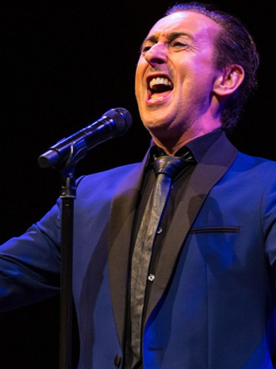 a white man in a blue jacket sings before a microphone his arms outspread