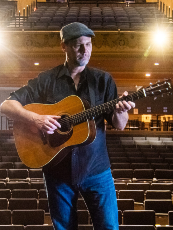 a white man in a cap plays an acoustic guitar on stage, an empty auditorium at his back