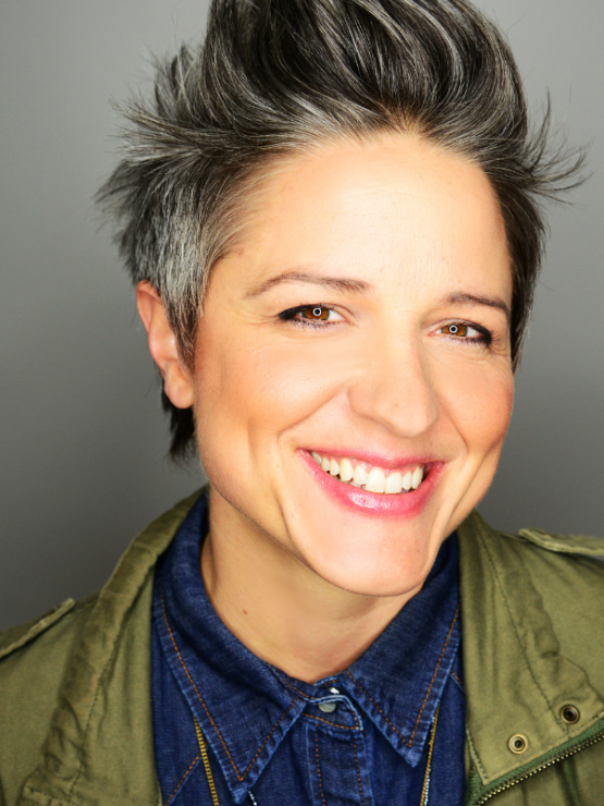 a woman with short salt-and-pepper hair swept up in a vertical style. she wears a denim shirt and an olive green jacket and smiles brilliantly