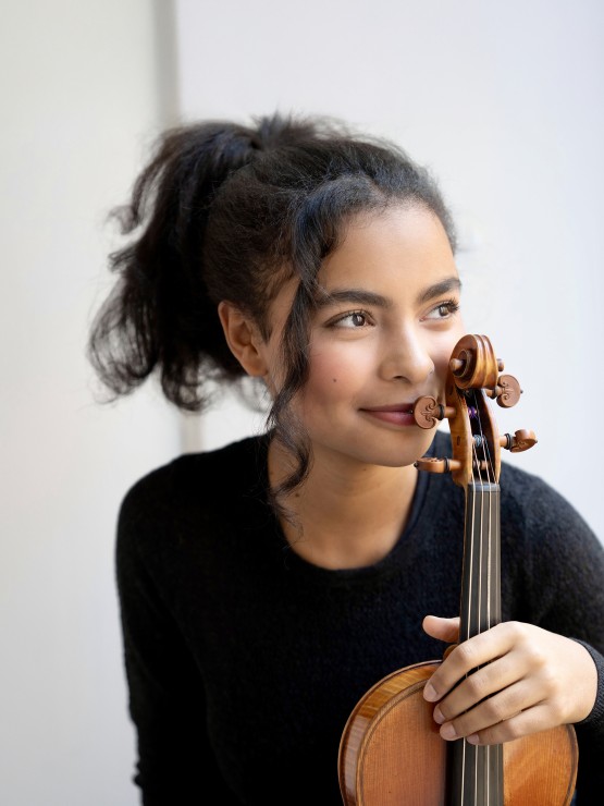 a violinist in her late teens holds the scroll of her violin to her cheek, looking to the side with a neutral smile