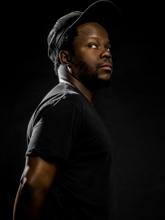 Ambrose Akinmusire, a black man in a black shirt and hat in front of a black background. 