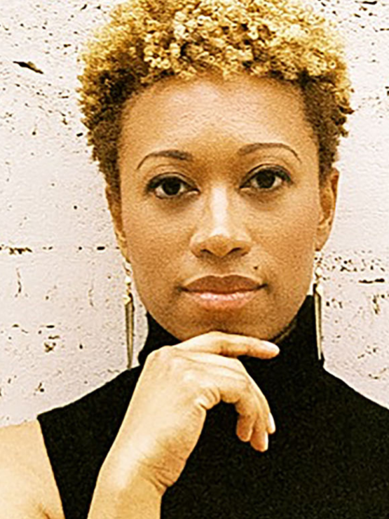 a black woman with short in a mock-necked black sleeveless shirt, against a painted concrete wall. she raises one elegant hand to her chin, and her expression is thoughtful and appraising
