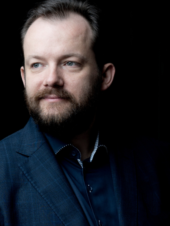 a bearded white man in his forties in a dark shirt and dark blue jacket, in a three-quarter view