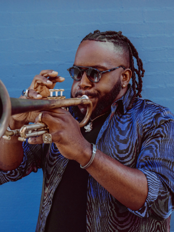in the foreground the bell of a trumpet. the trumpeter, Marquis Hill, is a black man with hair in twists on the top and a beard. He wears sunglasses and a blue shirt and stands in front of a blue wall