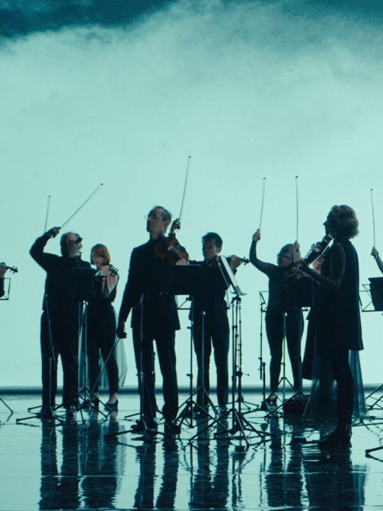 silhouetted before an aqua backdrop, a standing chamber orchestra raises their bows with a flourish