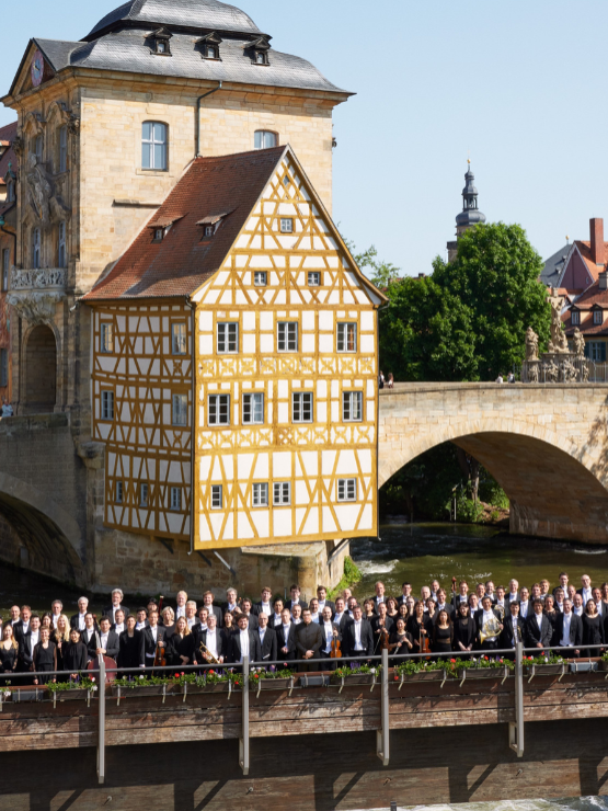 an orchestra lined up across a bridge with a yellow half-timbered building in the background