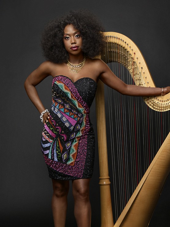 a black woman in her 30s, one hand on her golden harp and the other on her hip. She wears a multicolored iridescent strapless dress, and has voluminous shoulder-length curls
