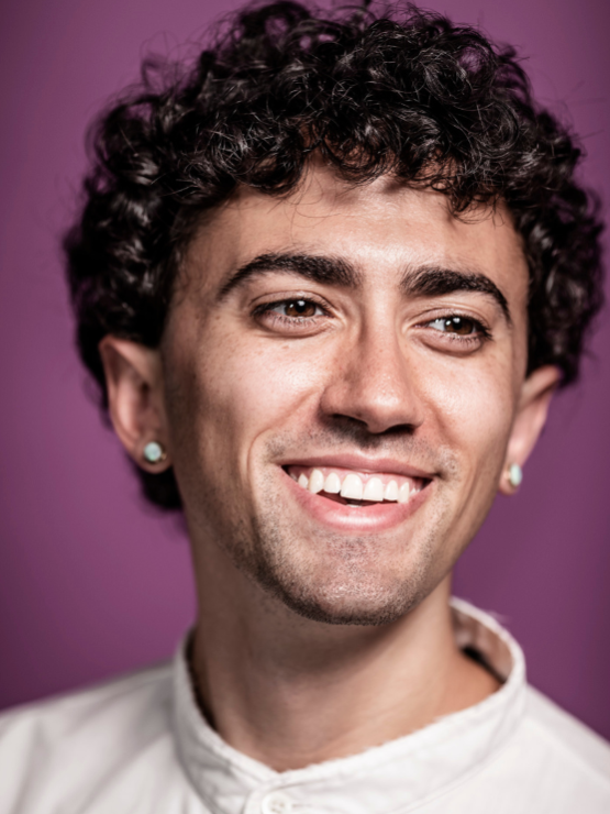 a white person in their late 20s with short brown curly hair, an ivory crewneck sweater, and stud earrings smiles in front of a purple background