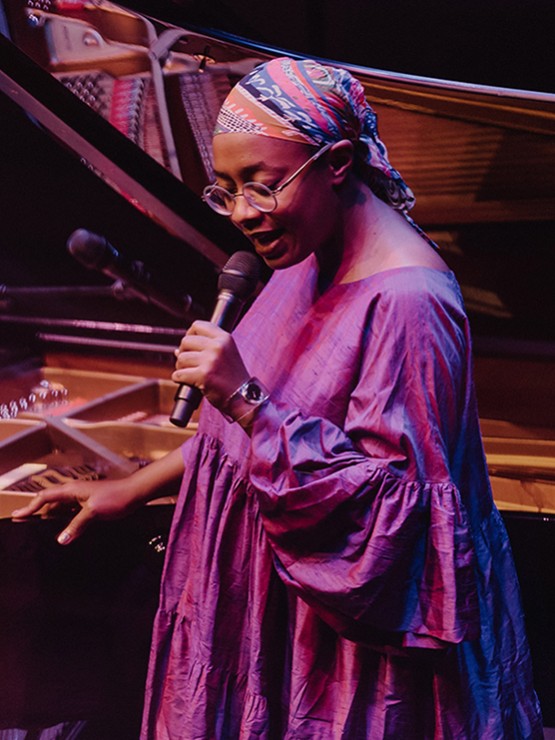 a black woman with glasses in a flowing purple robe, with a headwrap, sings in a concert, one hand on the rim of an open piano