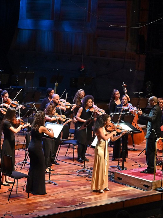 an orchestra with ethnically diverse members, standing, perform. A violin soloist, a woman in a long yellow dress, plays at the front.