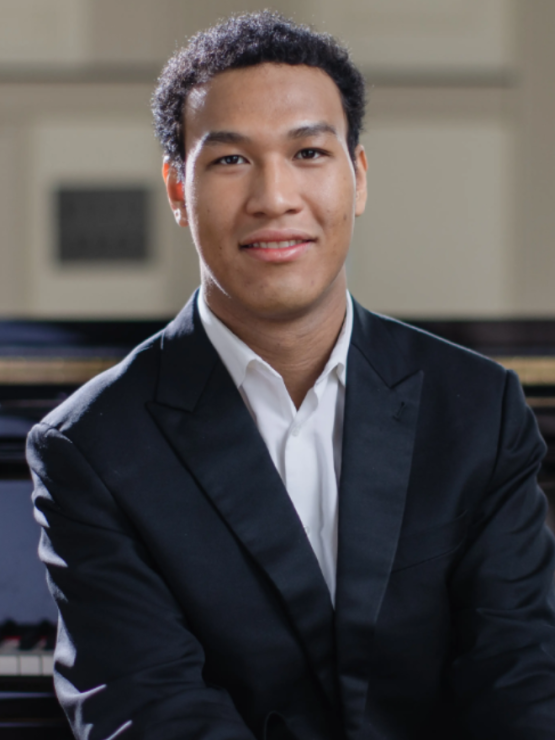 a young man in his early 20s with short dark curly hair and light brown skin sits in front of a grand piano in a brightly lit music room