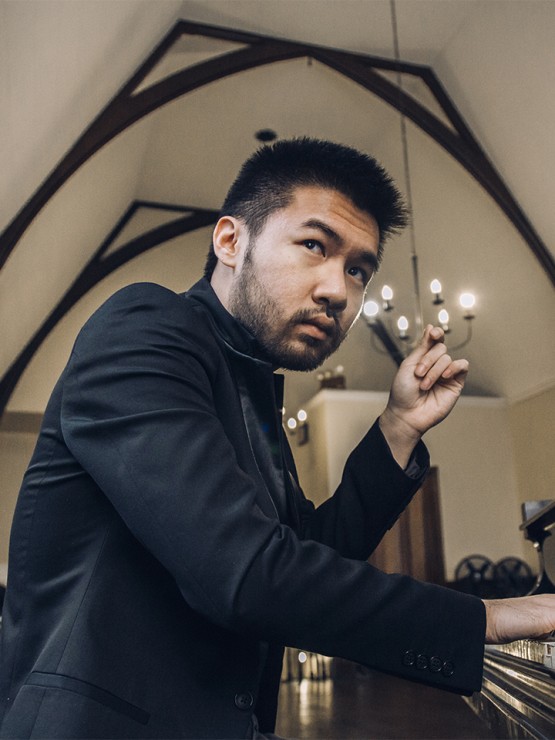 a young chinese-american man in a dark jacket plays the piano in a room with a vaulted ceiling. he has a scruffy short beard and one hand is lifted thoughtfully.