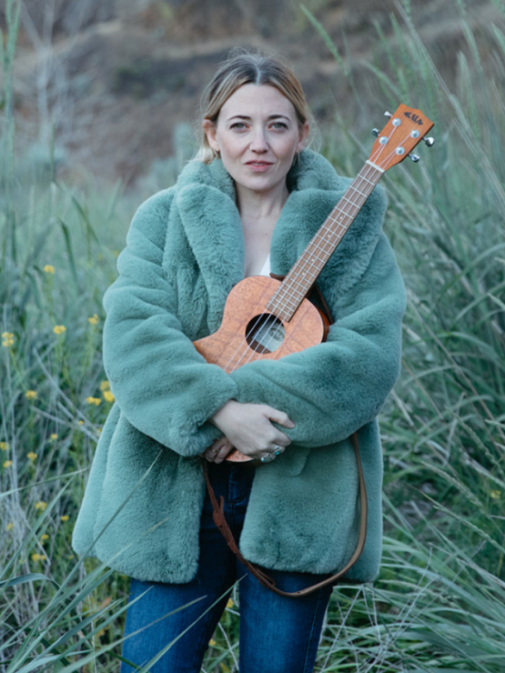a blonde woman in a fuzzy green coat holds an ukulele in a field amid tufts of grass