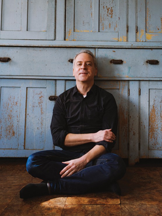 A white man in dark jeans and a black button-down shirt with the sleeves rolled up leans back against a rustic cupboard with flaking blue paint. 