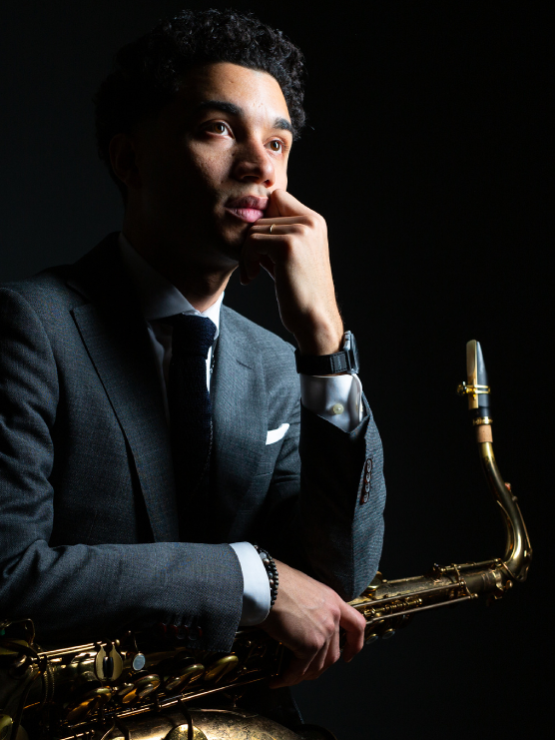 a puerto rican man with black hair wears a gray suit and faces right toward a light source. He holds a saxophone