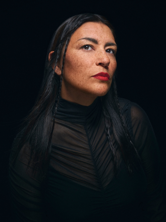 an indigenous woman in her 40s in black, against a dark background, in a three-quarter view. Her freckled face is framed by small braids, and she wears vivid red lipstick