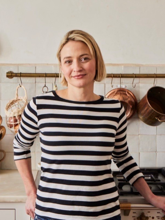 a blonde white woman with short hair and a black and white striped top stands in a bright kitchen