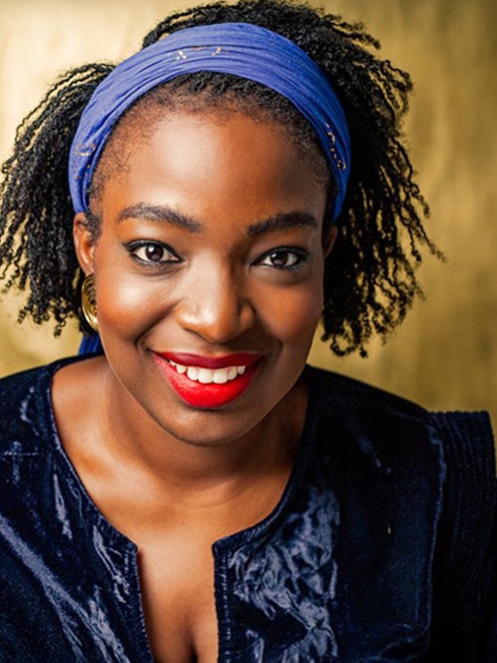 a warmly smiling black woman with a wide light blue headband. The lighting is golden, and she wears a chic navy jacket with a bright red lipstick