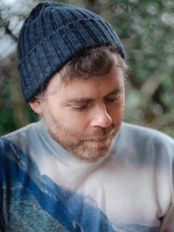 a white man with a short beard in his 30s wears a beanie and looks down