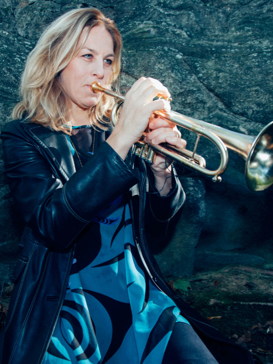 a blond white woman plays a trumpet outdoors in front of a large boulder