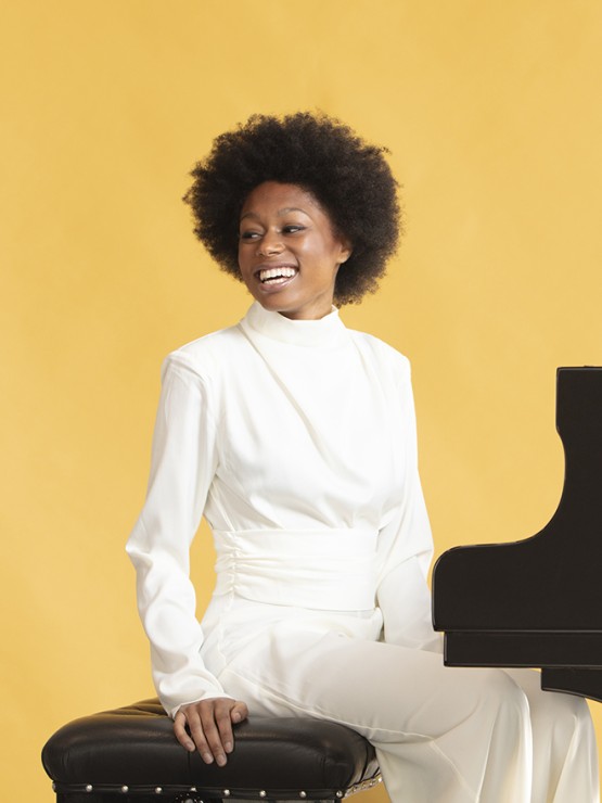 a young Black woman with a cheerful smile looks over her right shoulder as she sits on the bench of a grand piano. she wears an ivory sweater and trousers and the background is a sunny yellow