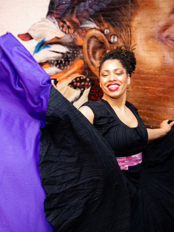 a multiracial black woman in a black dress swirls her skirt, revealing a wide purple hem. she stands in front of a mural depicting a brown-skinned elder whose face we cannot see