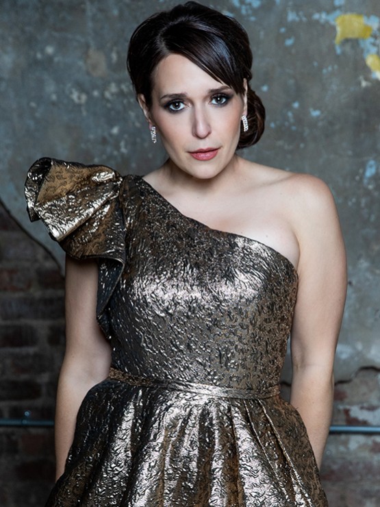 a woman with pale skin and dark hair wears a one-shoulder crinkly textured metallic fabric dress. her chin slightly tipped down, she makes intense eye contact.