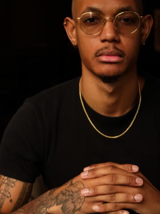 a young afro-dominican man with round gold-framed glasses and a shaved head. his arms are covered in detailed tattoos depicting music and wing motifs, and his elegant hands are folded in front of his chest.