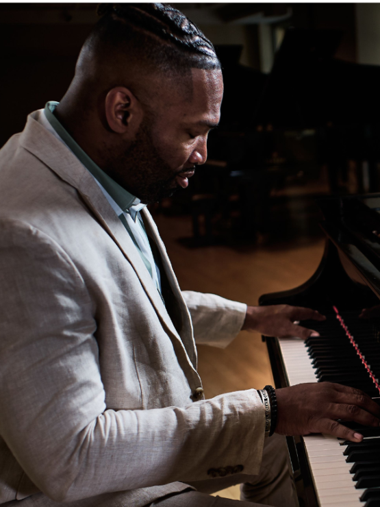 a black man in a light gray suit looks down at his hands as he sits sideways at a black piano