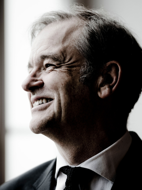 a white english man in his sixties turns to the side, smiling. the lighting is bright but cool, and he wears a jacket and tie