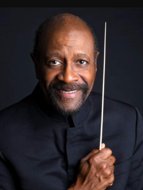 a black man in his 60s, with a goatee and a bald head, wears a black turtleneck against a black background. he holds a white conductor's baton vertically, close to the side of his face