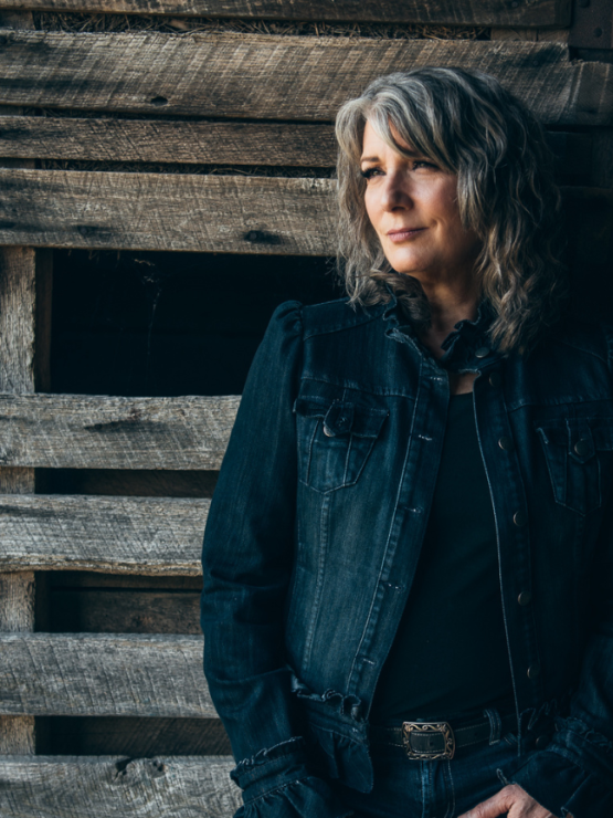a white woman with wavy gray hair stands in front of a rustic wall. She wears a dark ruffled denim jacket and dark jeans with a scrolled belt buckle.