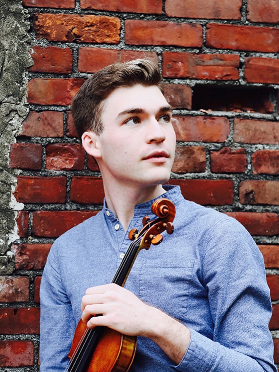 a young white man holds a violin and looks off and to his left. he stands outdoors in front of a brick wall.