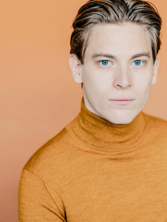a young white man with sandy hair and blue eyes wears an ochre turtleneck in front of a peach studio background