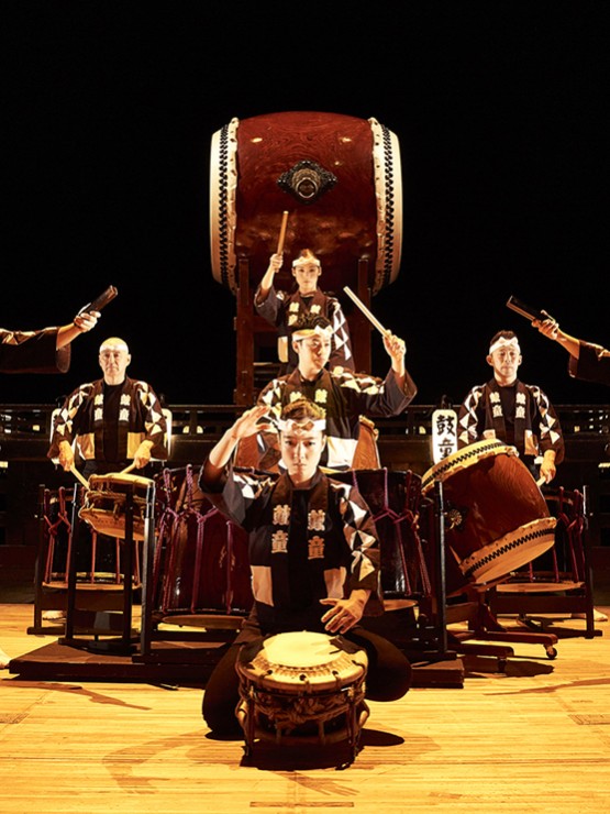 Eleven KODŌ drummers in black and white outfits gather on stage with their instruments.