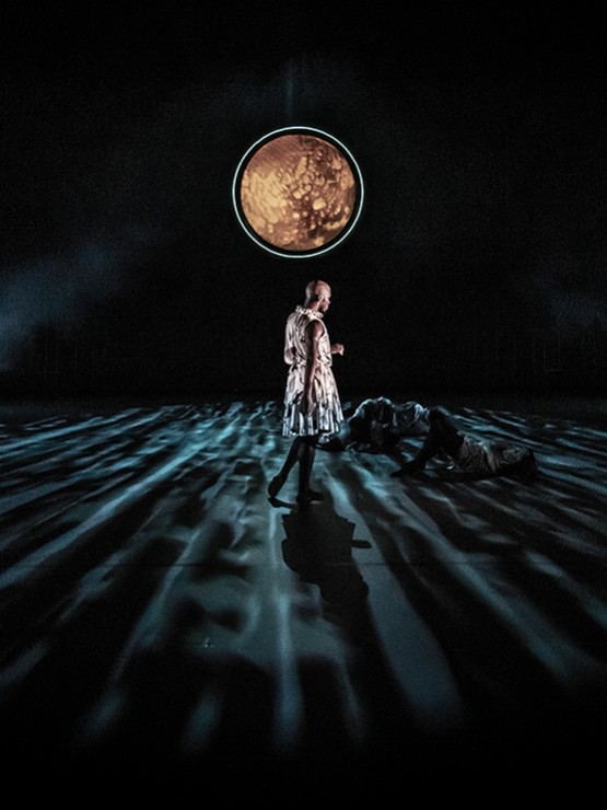 a male dancer stands alone on a dark stage, turned away from us toward two prone dancers and a moon-like illuminated circle