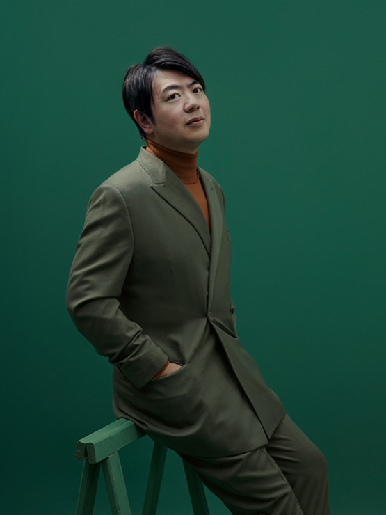 a chinese man in his early 40s wears an olive green suit with a russet turtleneck. he leans against a green sawhorse with his hands in his pockets, looking to his right toward us