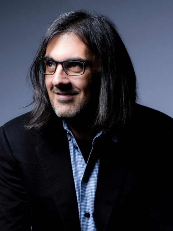 a greek man with shoulder length straight hair and glasses looks to his right. he wears a jacket and a light blue button down