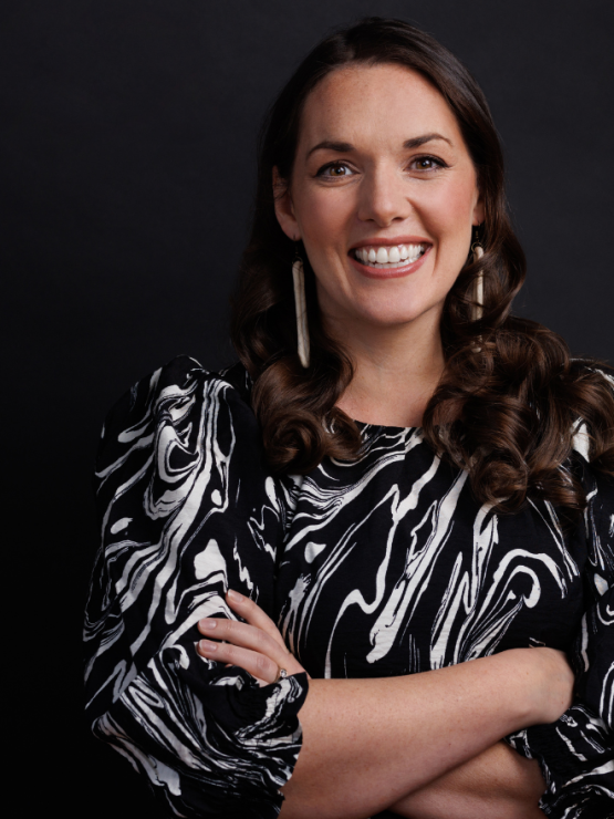 a white woman with shoulder length brown hair stands with her arms folded. she smiles and wears a black and white top.