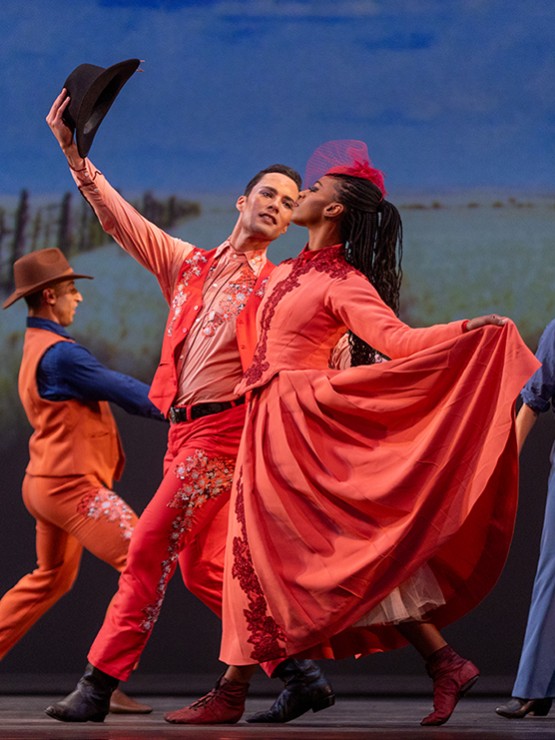 in a dance performance, a black woman in a full-skirted red western-style dress walks alongside a white man dressed as a cowboy in a red outfit