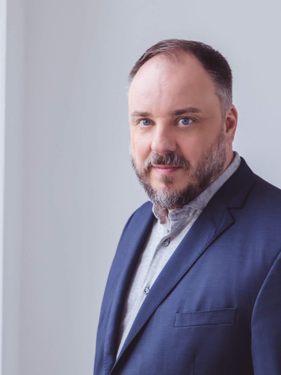 a german man with short graying hair and beard wears a light blue shirt and navy blue jacket. he turns slightly toward us