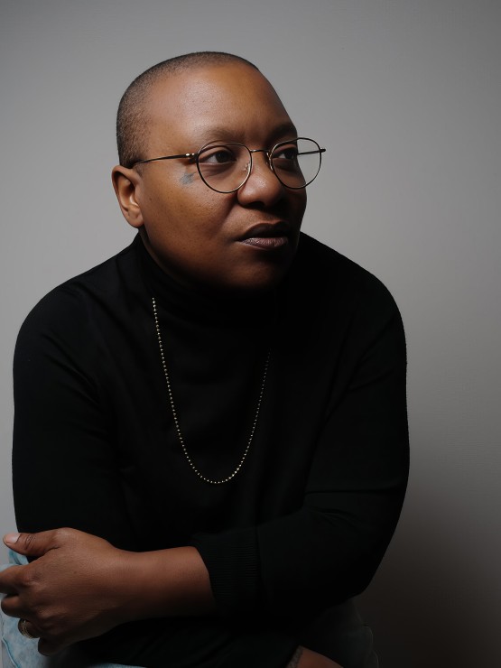 a black woman with a shaved head and thin framed round glasses. She sits with her arms around herself, leaning slightly forward, looking at us from the side