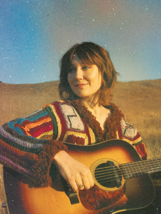 a young white woman plays the guitar in a striped sweater outside in an open field. The photo has a hazy vintage effect.