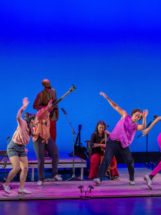 A multicultural group of tap dancers and musicians perform on low platforms