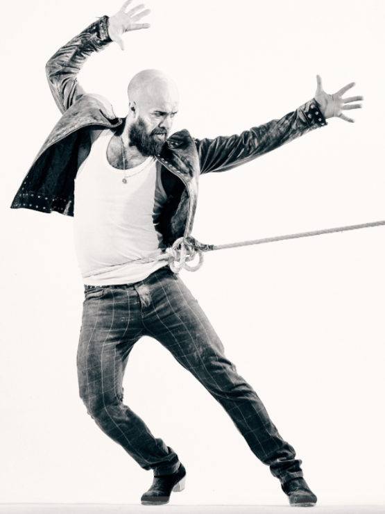 a bald-headed spanish man in heeled boots, jeans, and a leather jacket is tied with a rope around his waist. he leans away from the rope, arms raised